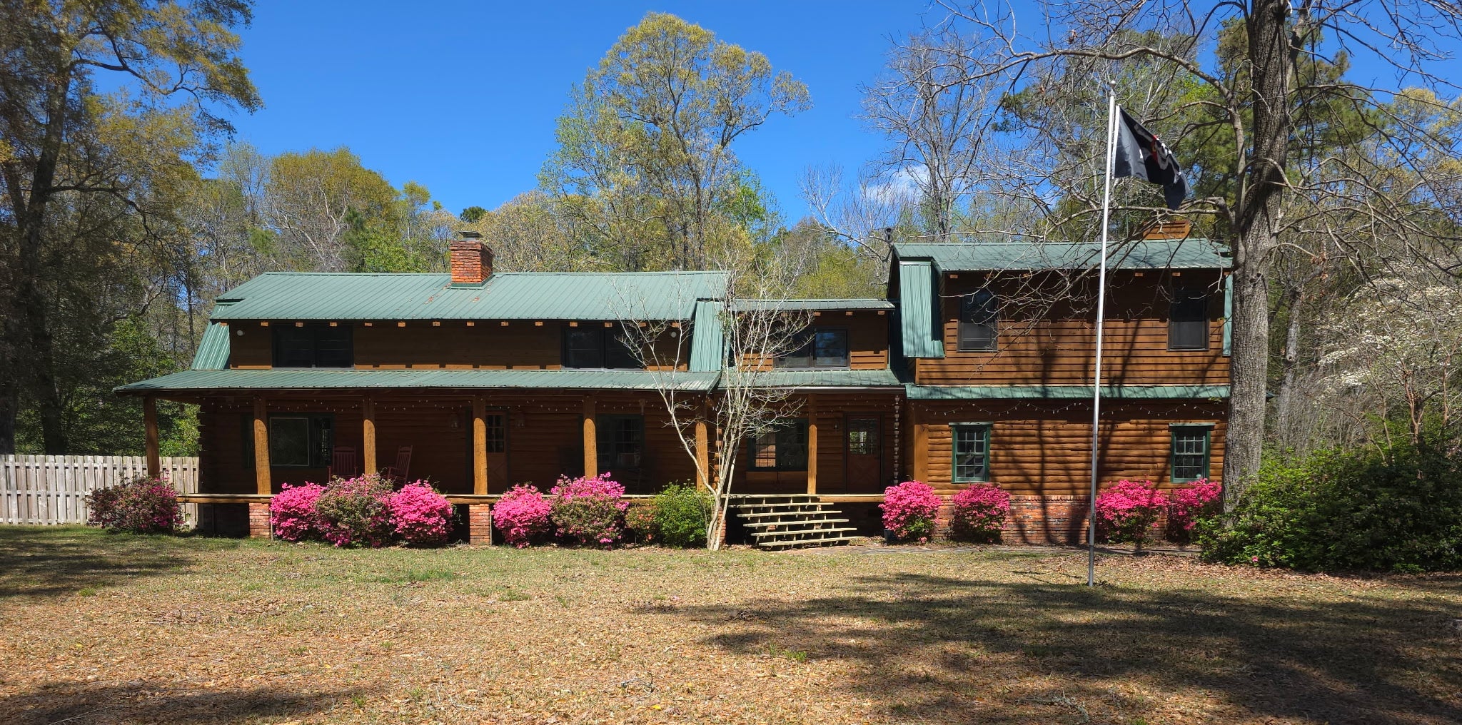 Wooden cabin with pink flowering bushes in front, surrounded by trees and clear blue sky.